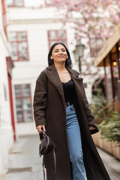 Cheerful woman in stylish coat and wireless earphones walking on urban street in prague — Stock Photo