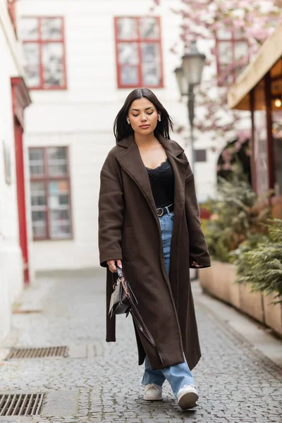 Piena lunghezza di elegante donna bruna in auricolari wireless a piedi sulla strada stretta a Praga — Foto stock