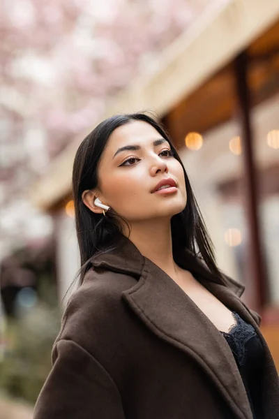 Young brunette woman in brown coat looking away while listening music in wireless earphone on street in prague — Stock Photo