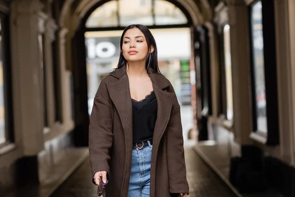 Stylish woman in brown coat and jeans looking away near arch building in prague — Stock Photo