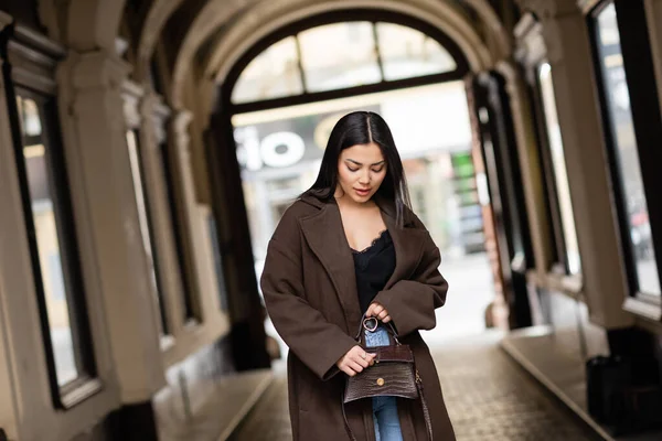 Young woman in autumn coat opening handbag near arch building on blurred background in prague — Stock Photo