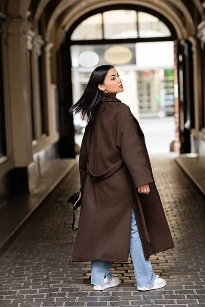 Rückansicht einer jungen brünetten Frau in braunem Mantel, die unter einem Bogengebäude in Prag wegschaut — Stockfoto