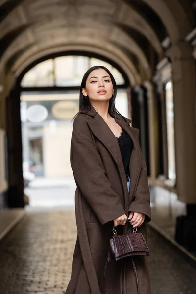 Молода жінка в модному осінньому пальто тримає сумку і дивиться на вулицю в празі — стокове фото