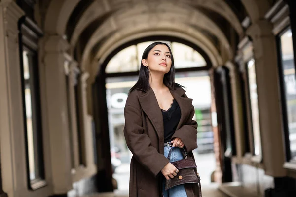 Morena mujer en abrigo sosteniendo el bolso y mirando hacia otro lado cerca de arco edificio en praga - foto de stock