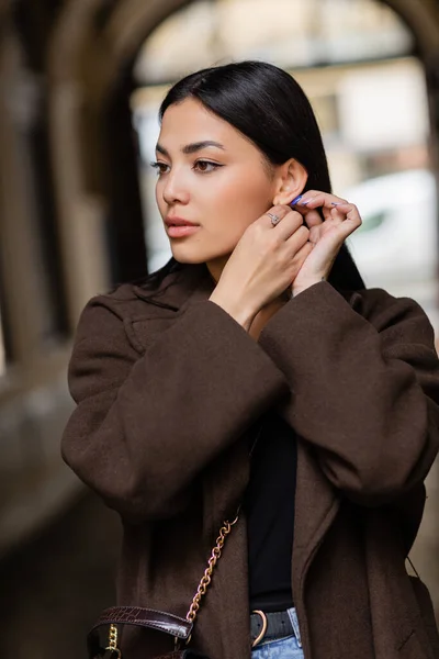 Brunette woman in autumn coat adjusting earring on blurred street in prague — Stock Photo