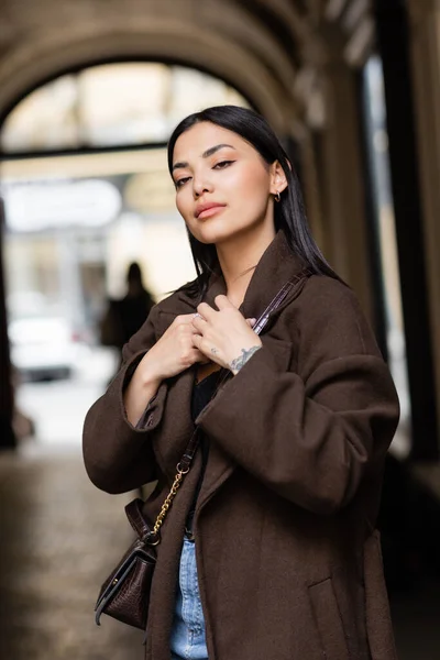 Giovane donna bruna in cappotto marrone alla moda con crossbody su sfondo sfocato a praga — Foto stock
