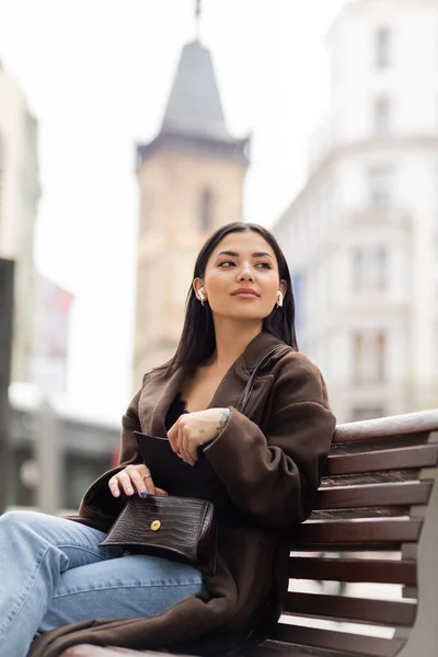 Jovem mulher em fones de ouvido sem fio abrindo bolsa da moda e olhando para longe no banco em prague — Fotografia de Stock