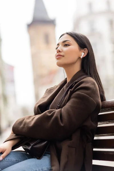 Brunette femme en manteau marron écouter de la musique dans écouteur sans fil sur banc dans la prague — Photo de stock