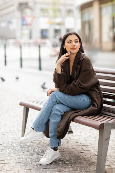 Hübsche Frau in trendiger Herbstkleidung hört Musik in drahtlosen Kopfhörern auf Bank in Prag und schaut weg — Stockfoto
