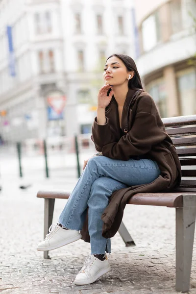 Mujer soñadora sentada en el banco en prague y escuchando música en auriculares inalámbricos - foto de stock