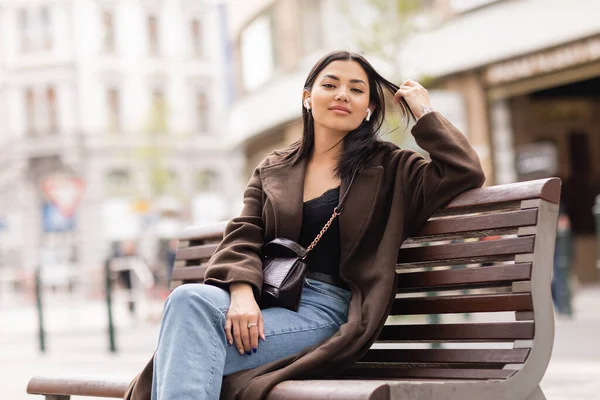 Happy brunette woman touching hair and listening music in wireless earphones on bench in prague — Stock Photo