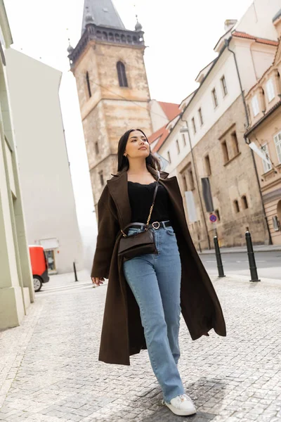 Brunette woman in brown coat and jeans walking along ancient street in prague — Stock Photo