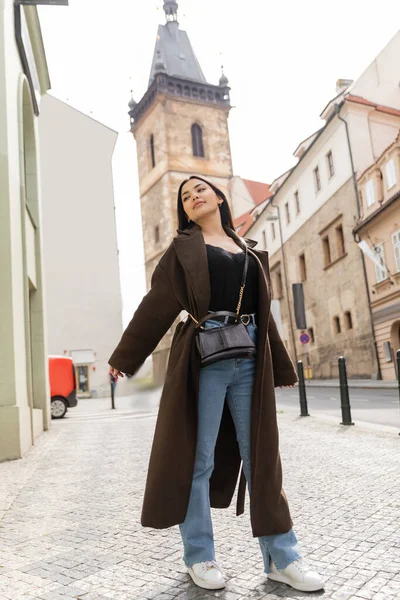 Улыбающаяся женщина в джинсах и коричневом пальто стоящая с протянутыми руками на древней улице в Праге — стоковое фото