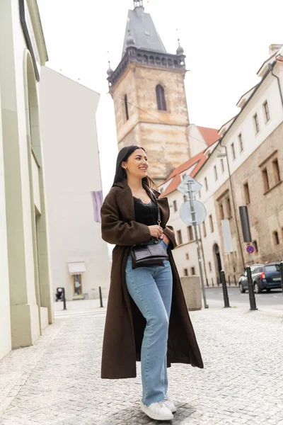 Comprimento total de mulher morena feliz em casaco da moda e jeans de pé na rua antiga em prague — Fotografia de Stock