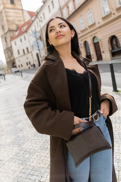 Young woman in trendy coat standing with hands in pockets of jeans and looking away in prague — Stock Photo