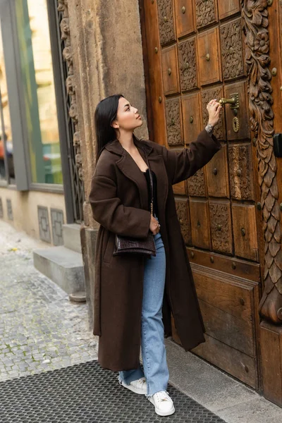 Comprimento total da mulher morena em casaco elegante com abertura crossbody esculpida porta de madeira do edifício em prague — Fotografia de Stock