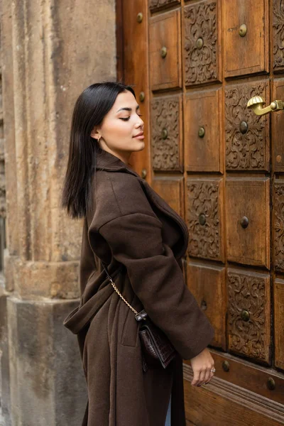 Mulher morena vestindo casaco marrom na moda com crossbody e pé perto de porta de madeira esculpida em prague — Fotografia de Stock