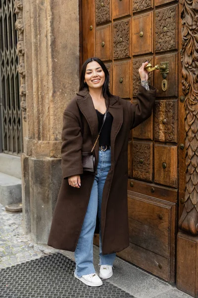 Mulher morena feliz no casaco da moda tocando maçaneta da porta perto do edifício em prague — Fotografia de Stock