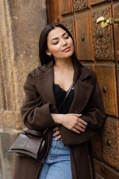 Positive brunette woman in stylish autumn coat standing near carved wooden door in prague — Stock Photo
