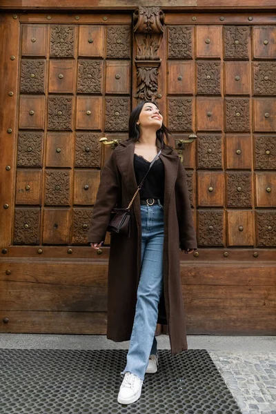 Full length of happy woman in jeans and coat looking away near carved door on street in prague — Stock Photo
