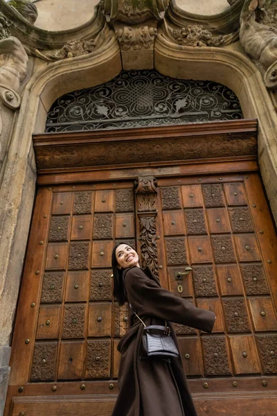 Low angle view of cheerful woman in trendy coat with crossbody near wooden carved door in prague — Stock Photo