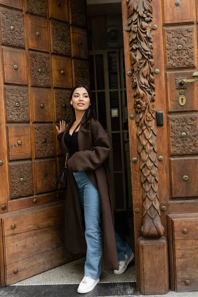 Full length of brunette woman in stylish coat looking away while opening wooden door in prague — Stock Photo