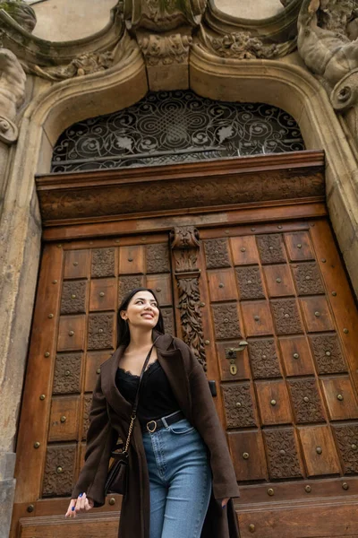 Low angle view of brunette woman in jeans and trendy coat smiling near carved wooden door in prague — Stock Photo