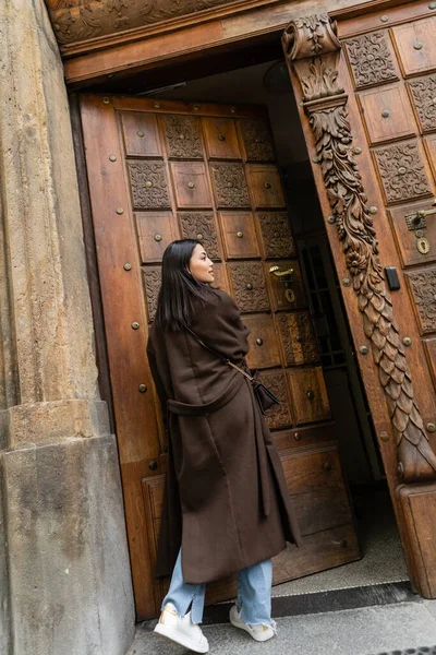 Joven morena en abrigo marrón caminando en puerta de madera abierta en prague - foto de stock