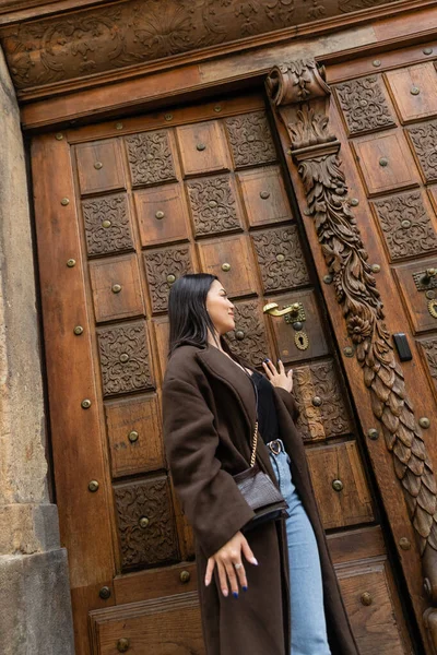 Joven morena mujer en abrigo marrón apertura de madera tallada puerta en prague - foto de stock