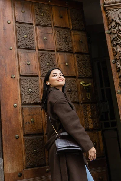 Morena mujer con abrigo y auricular inalámbrico sonriendo cerca de la puerta abierta tallada en praga - foto de stock