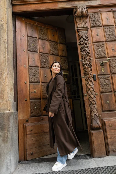 Comprimento total da mulher elegante no casaco marrom sorrindo e olhando para longe perto da porta de madeira em prague — Fotografia de Stock
