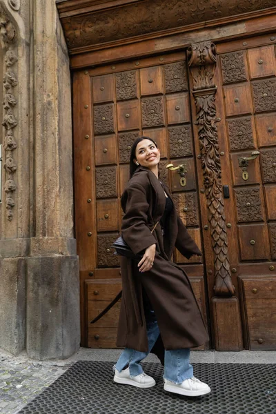 Mujer alegre en el abrigo marrón de moda caminando cerca de la puerta de madera tallada en praga y mirando hacia otro lado - foto de stock
