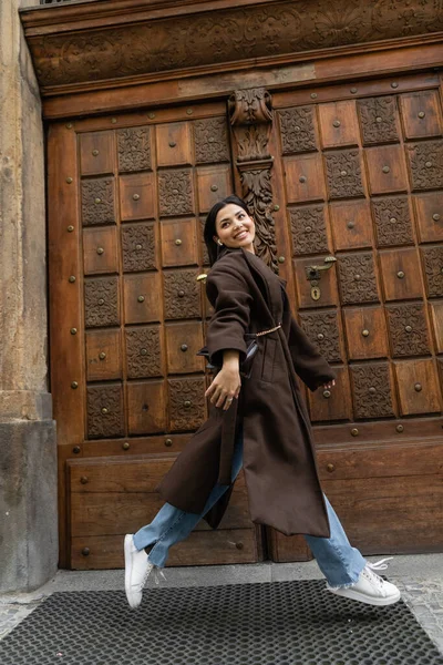 Pleine longueur de femme brune excitée en manteau élégant courir près de porte sculptée en bois sur la rue dans la prague — Photo de stock