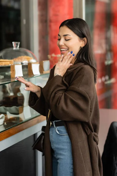 Femme étonnée tenant la main près du visage tout en regardant vitrine de pâtisserie boutique avec desserts à la prague — Photo de stock