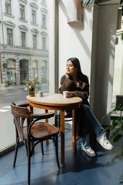 Young woman in autumn coat sitting in cafe near window and looking at street in prague — Stock Photo