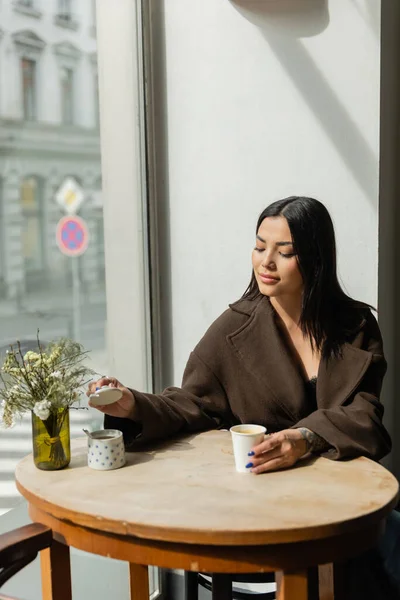 Femme brune avec tasse en papier ouvrant bol à sucre tout en étant assis près de la fenêtre dans un café prague — Photo de stock