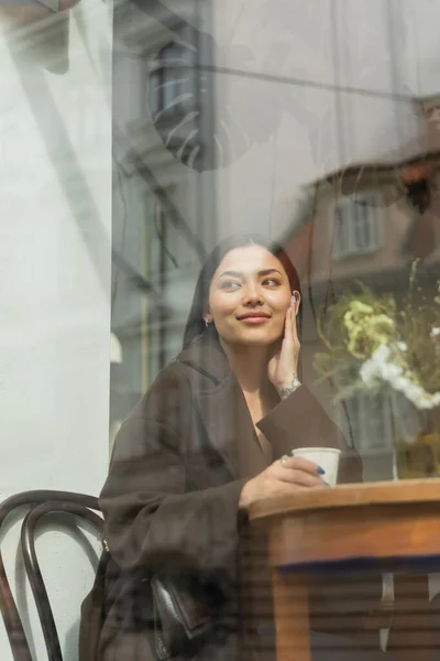 Femme heureuse regardant à l'extérieur tout en étant assis près de la fenêtre dans prague café — Photo de stock