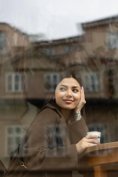 Улыбающаяся женщина в пальто держит бумажную чашку возле стекла в кафе — стоковое фото