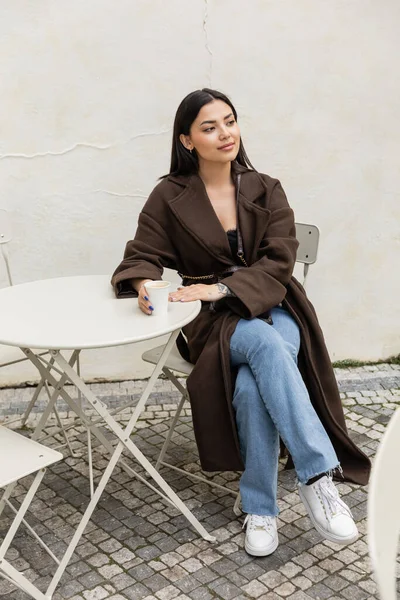 Junge brünette Frau im Mantel hält Pappbecher neben Tisch in einem Café in Prag — Stockfoto