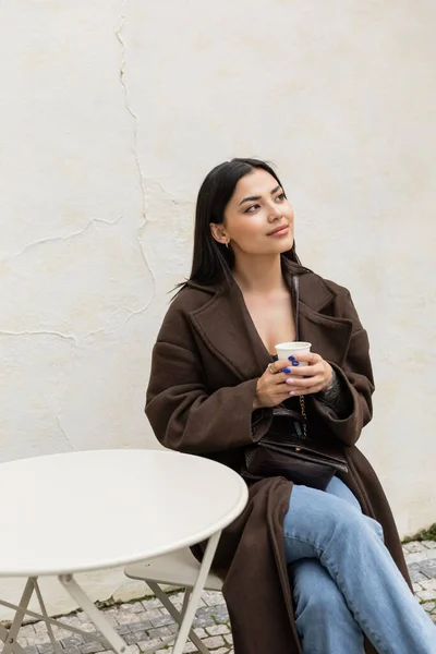 Bruna donna in cappotto in possesso di tazza di carta vicino edificio in caffè all'aperto a Praga — Foto stock