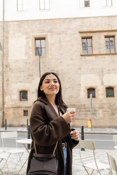 Радостная женщина в пальто держит бумажную чашку с кофе на городской улице в Праге — стоковое фото
