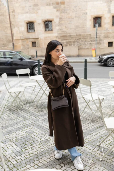 Полная длина стильной молодой женщины, пьющей кофе возле стульев открытого кафе на улице в Праге — стоковое фото