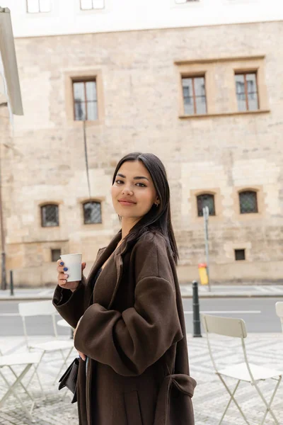 Porträt einer jungen Frau im Mantel, die Pappbecher in der Hand hält und auf der Straße in Prag in die Kamera lächelt — Stockfoto