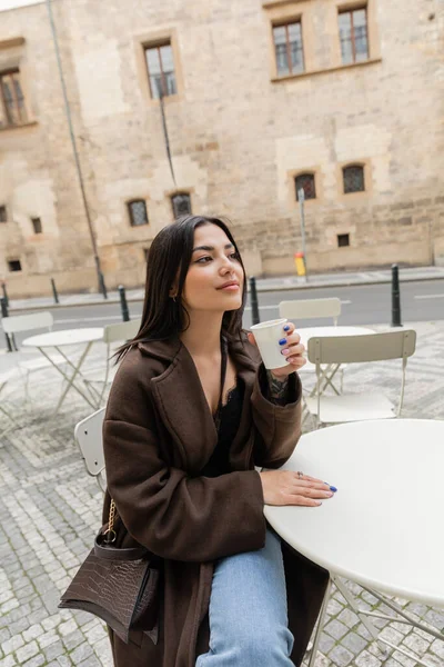 Mulher morena no casaco segurando copo de papel com café no café ao ar livre na rua em Praga — Fotografia de Stock