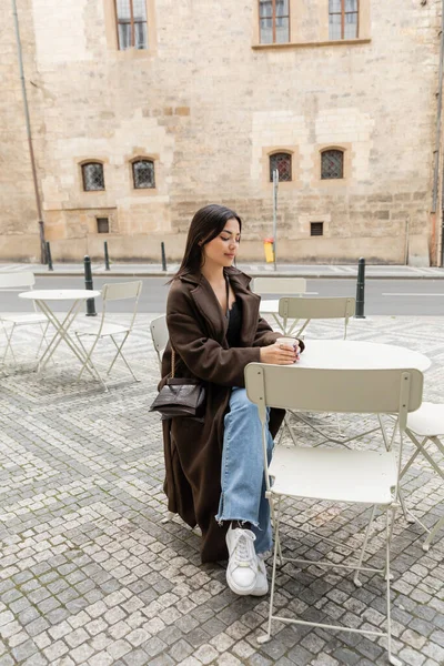 Turista na moda em casaco segurando copo de papel com café no café ao ar livre na rua em Praga — Fotografia de Stock