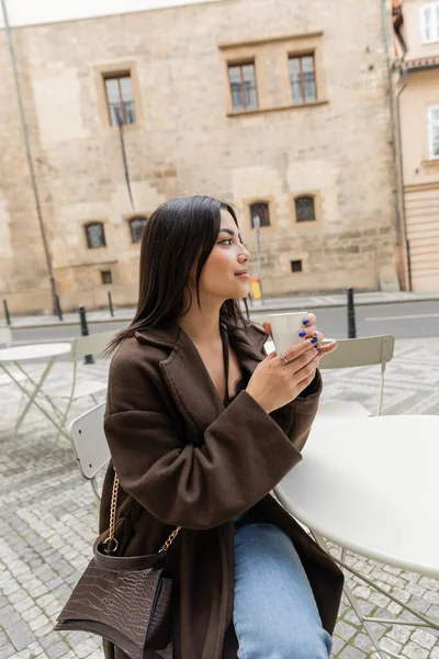 Вид сбоку на стильную молодую женщину в коричневом пальто, держащую бумажный стаканчик с кофе в открытом кафе в Нью-Йорке — стоковое фото