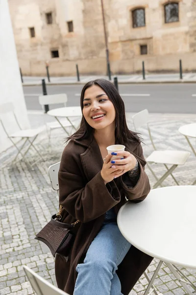 Touriste brune positive en manteau tenant tasse en papier avec café près de la table dans un café extérieur à Prague — Photo de stock