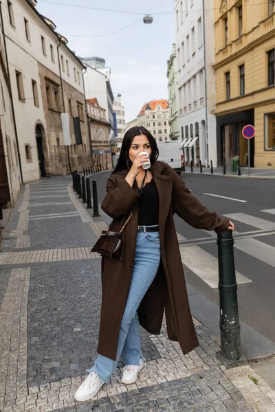 Stylish woman in coat drinking coffee from paper cup on street in Prague — Stock Photo