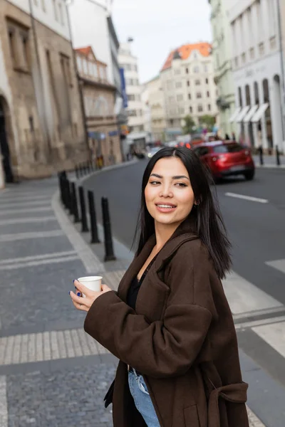 Portrait of stylish brunette tourist holding paper cup on blurred street in Prague — Stock Photo