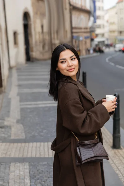 Brunette tourist in coat holding paper cup on urban street in Prague — Stock Photo